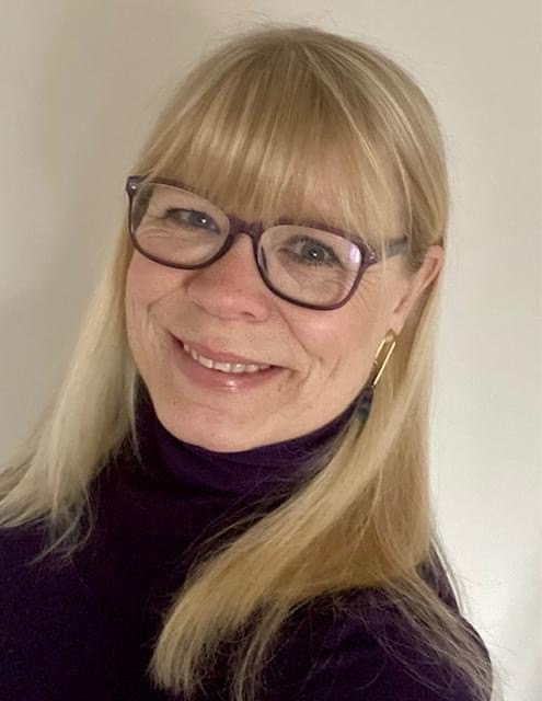 A woman with long blonde hair and glasses smiles at the camera, wearing a dark top and gold earrings.