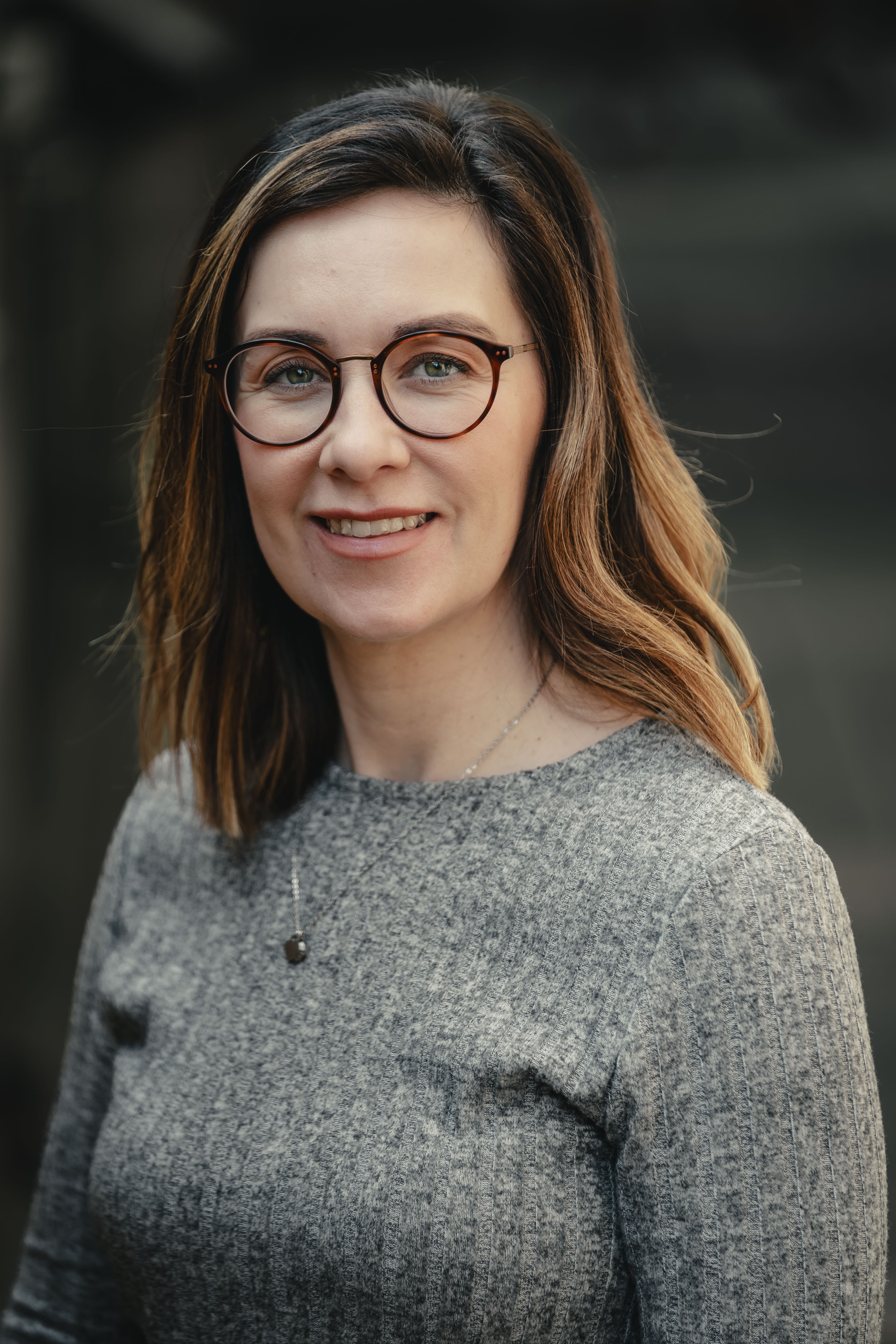 Portrait of a woman with shoulder-length brown hair wearing round glasses, a grey textured top, and a pendant necklace, smiling softly against a blurred background.