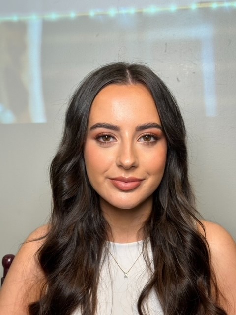 Portrait of a young woman with long, dark wavy hair, wearing a white top and subtle makeup, seated against a light grey background.