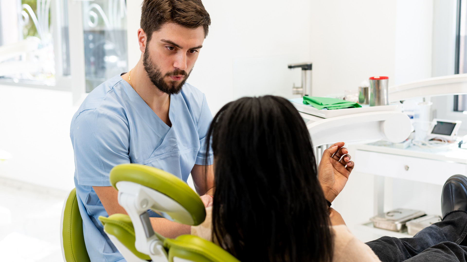 A dentist in blue scrubs speaks with a patient seated in a dental chair during a consultation in a modern clinical setting.