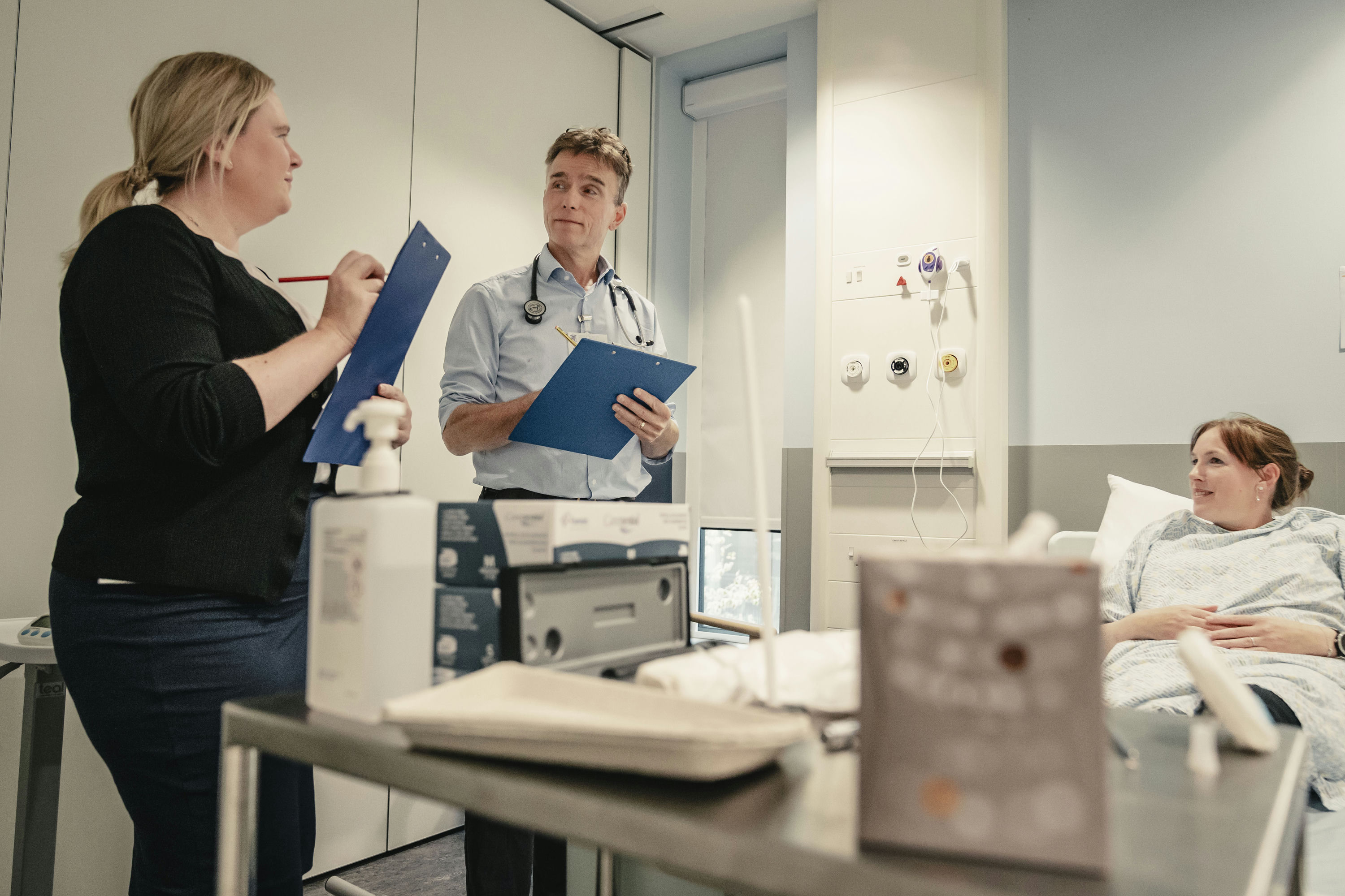 Two examiners with clipboards standing next to a patient in a hospital bed