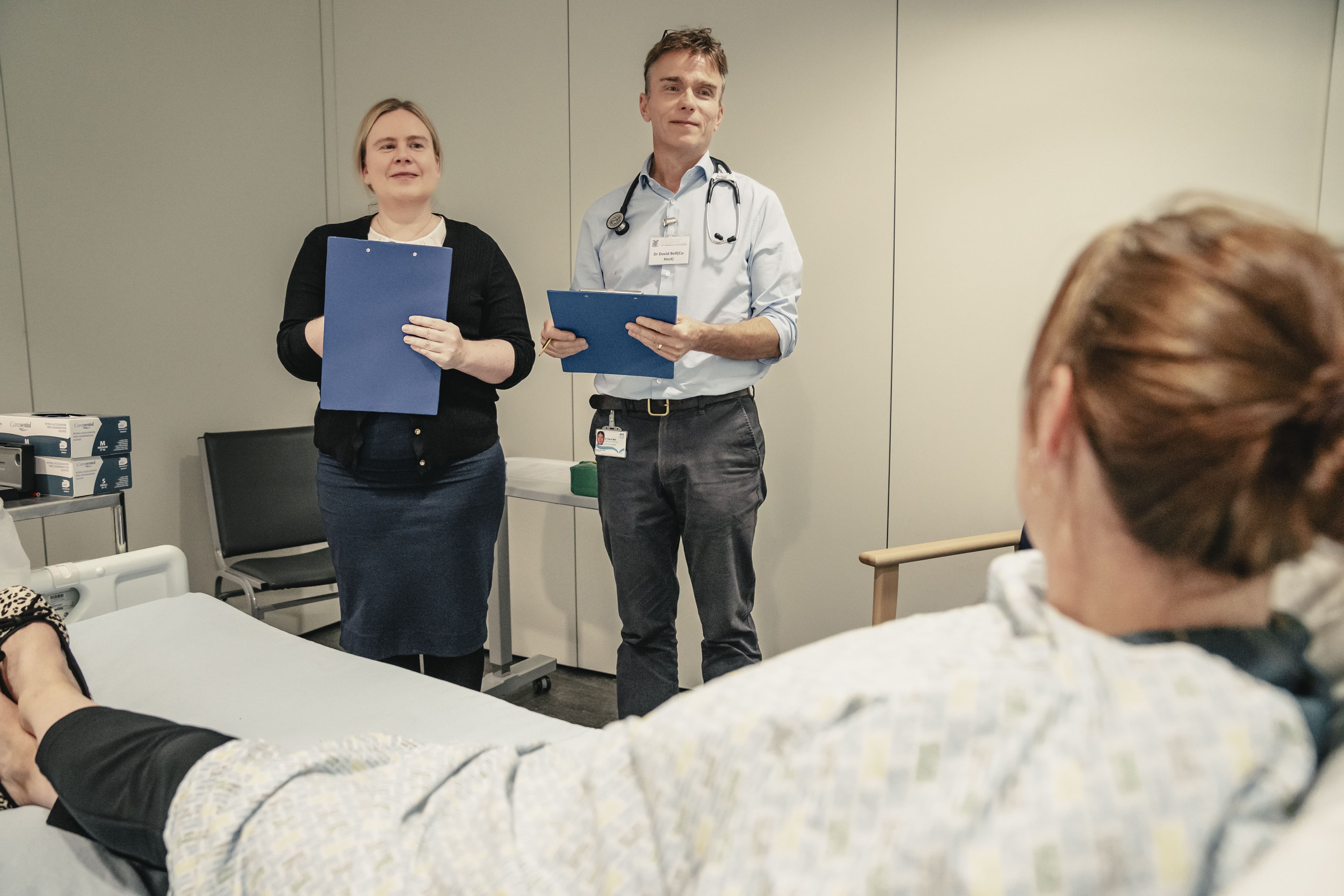 Two healthcare professionals with clipboards assess a patient lying in a hospital bed