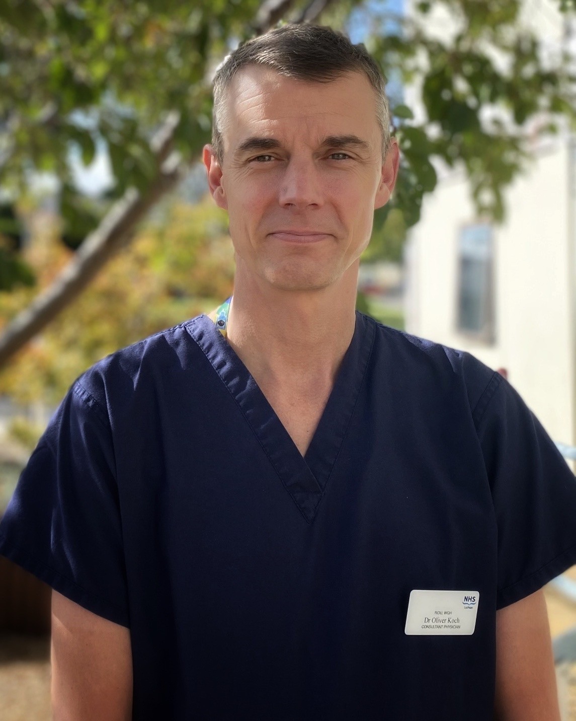 Dr Oliver Koch, Consultant Physician, wearing navy NHS scrubs and standing outdoors with trees in the background.