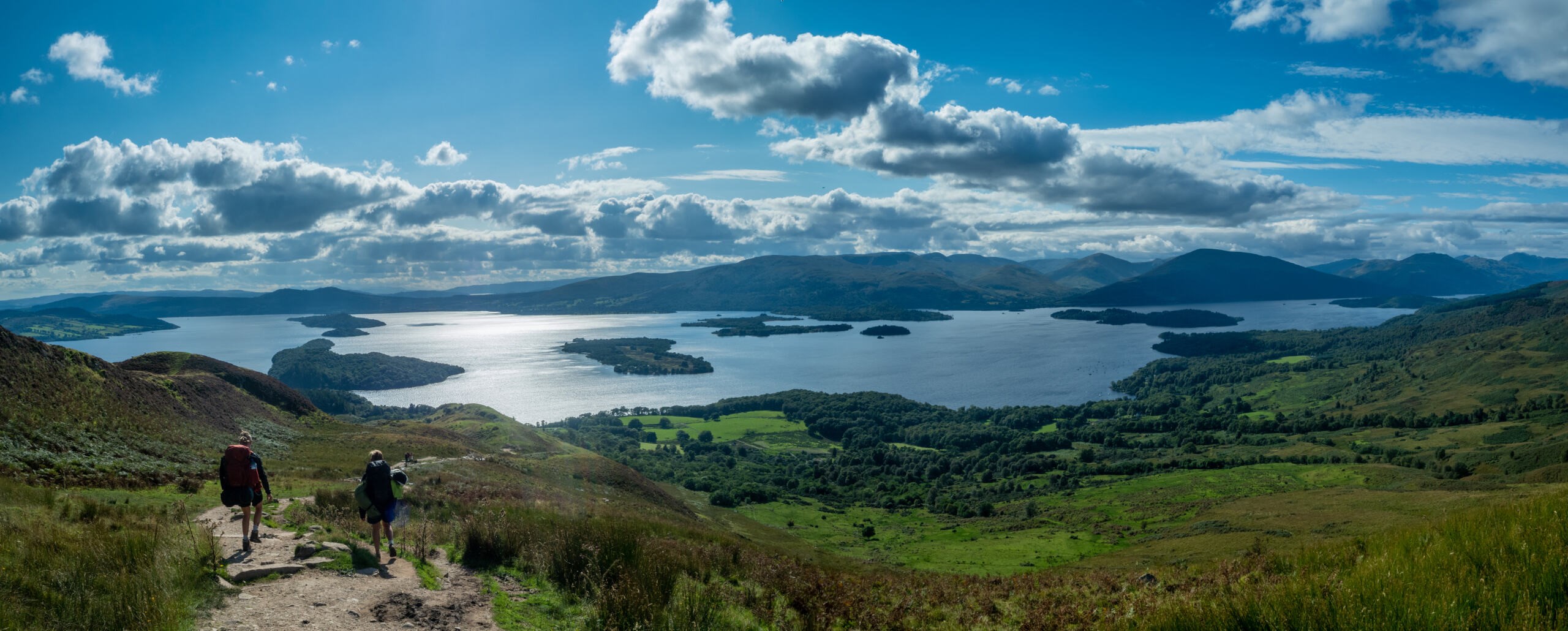 Two people walking down hill with rolling lush green landscape overlooking lake